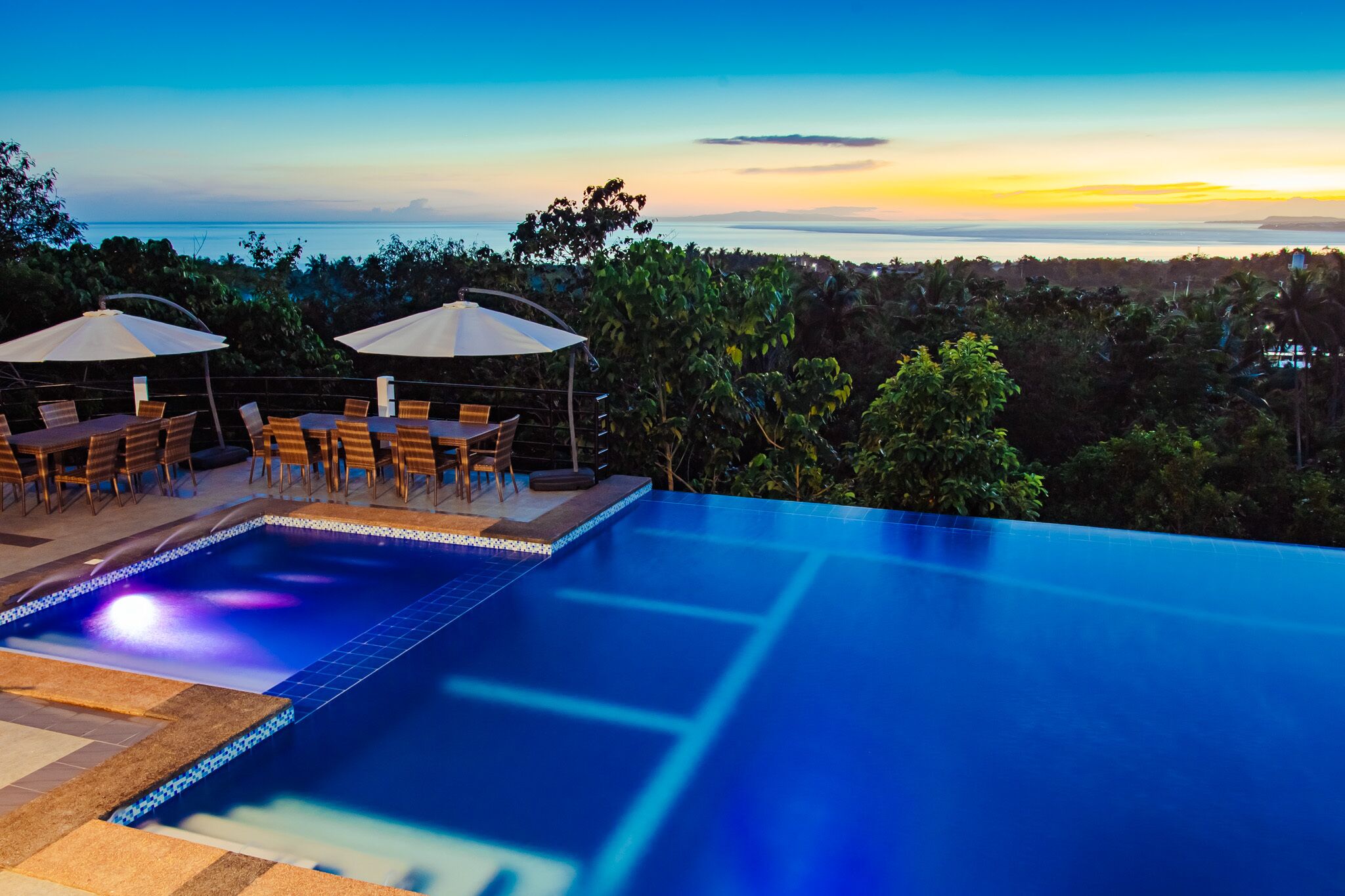 A swimming pool with a view of the ocean at sunset, surrounded by outdoor seating and umbrellas.