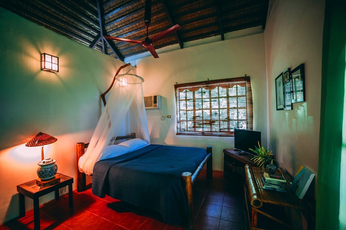 A room with a bed, a bedside lamp, a TV, a window with blinds, and a ceiling fan.