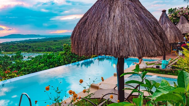 Una piscina infinita con vistas a un paisaje exuberante, rodeada de sombrillas de paja y sillones de descanso.