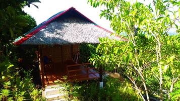 A thatched-roof hut with a wooden porch, surrounded by lush greenery.