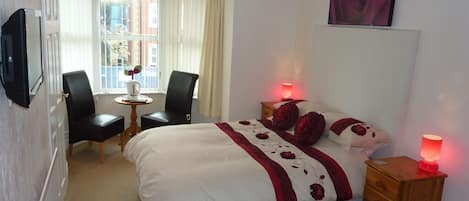 A hotel room with a bed, two chairs, a small table, a nightstand, a TV, and a window with curtains.