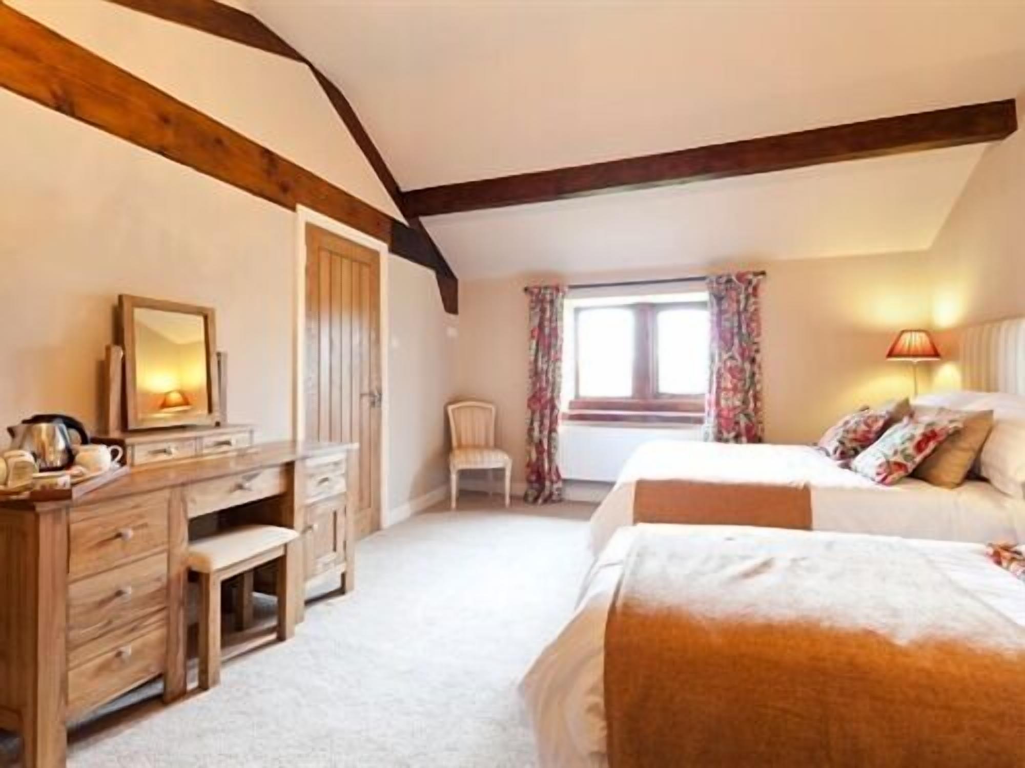 Een slaapkamer met een houten dressoir, een stoel, een bed met bloemenkussens, en een raam met gordijnen.