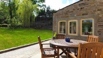Un bâtiment en pierre avec une table et des chaises sur une terrasse, entouré d’une pelouse et d’arbres.
