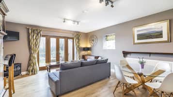 A modern living room with a grey sofa, a glass dining table with white chairs, a wooden floor, and a large window with curtains.