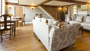 Amplia sala de estar con una escalera de madera, un comedor con mesa y sillas de madera, y un sofá grande con cojines estampados.