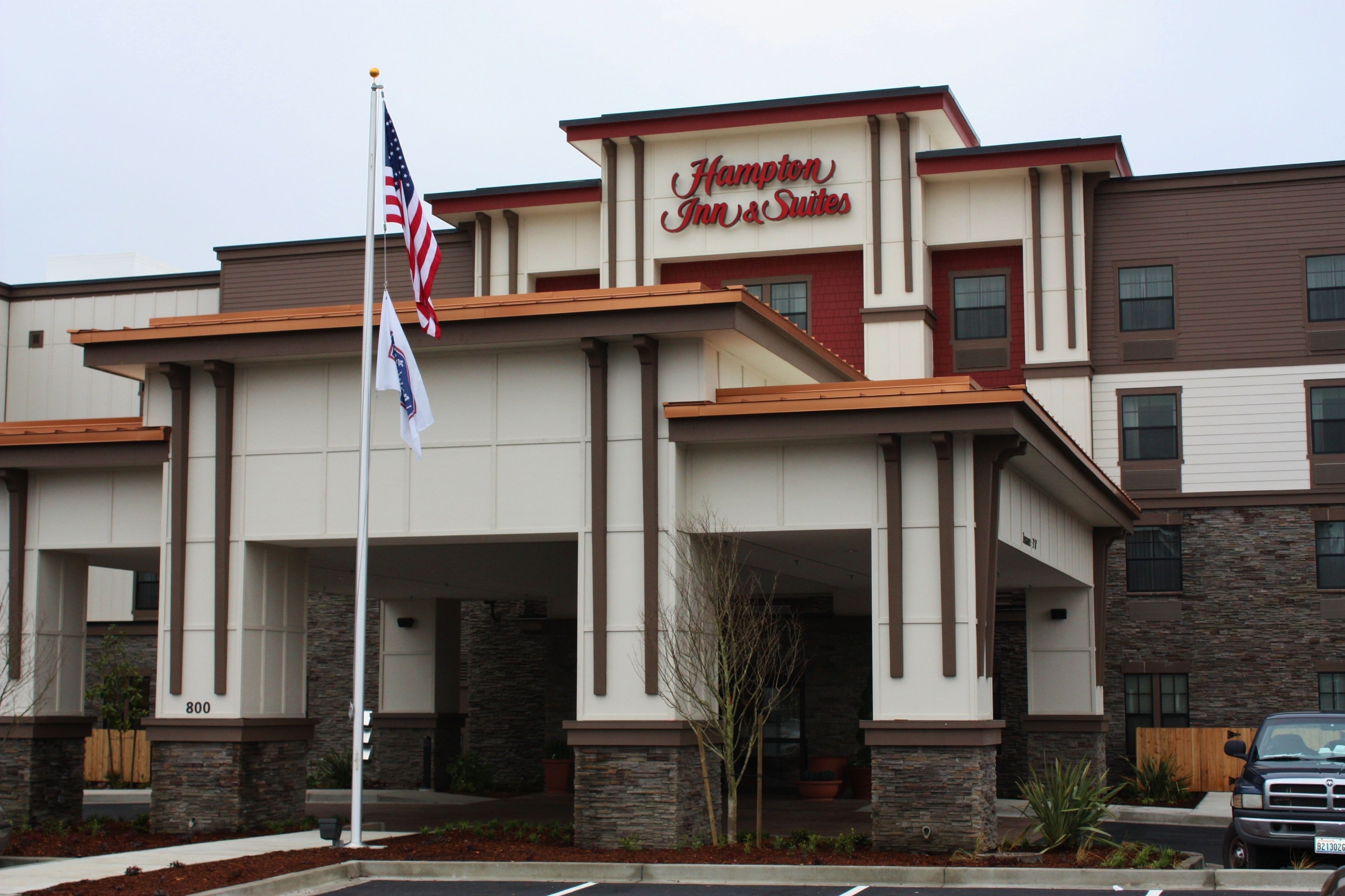 Un bâtiment de la chaîne Hampton Inn & Suites, doté d’une architecture moderne, avec une façade en pierre et un drapeau américain bien visible.