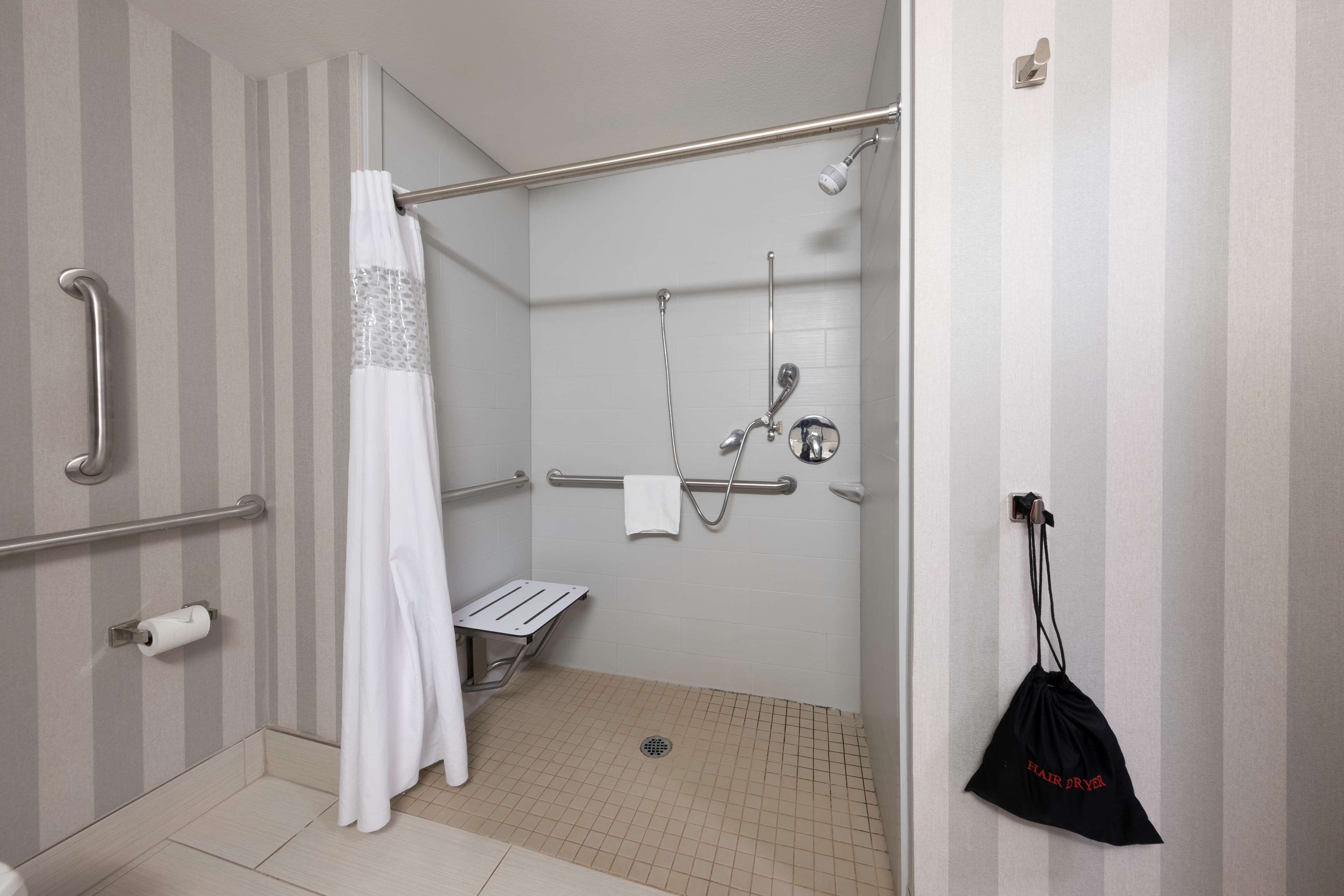 Área de chuveiro acessível para cadeirantes, com barras de apoio, chuveiro e porta-toalhas.