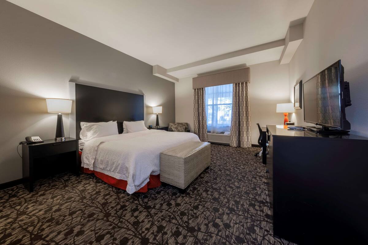 A hotel room with a large bed, bedside tables with lamps, a desk with a flat-screen TV, and a window with curtains.
