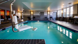 Une grande piscine intérieure à l’eau d’un bleu limpide, entourée de chaises longues et d’un espace baigné de soleil, avec des tables et des chaises.