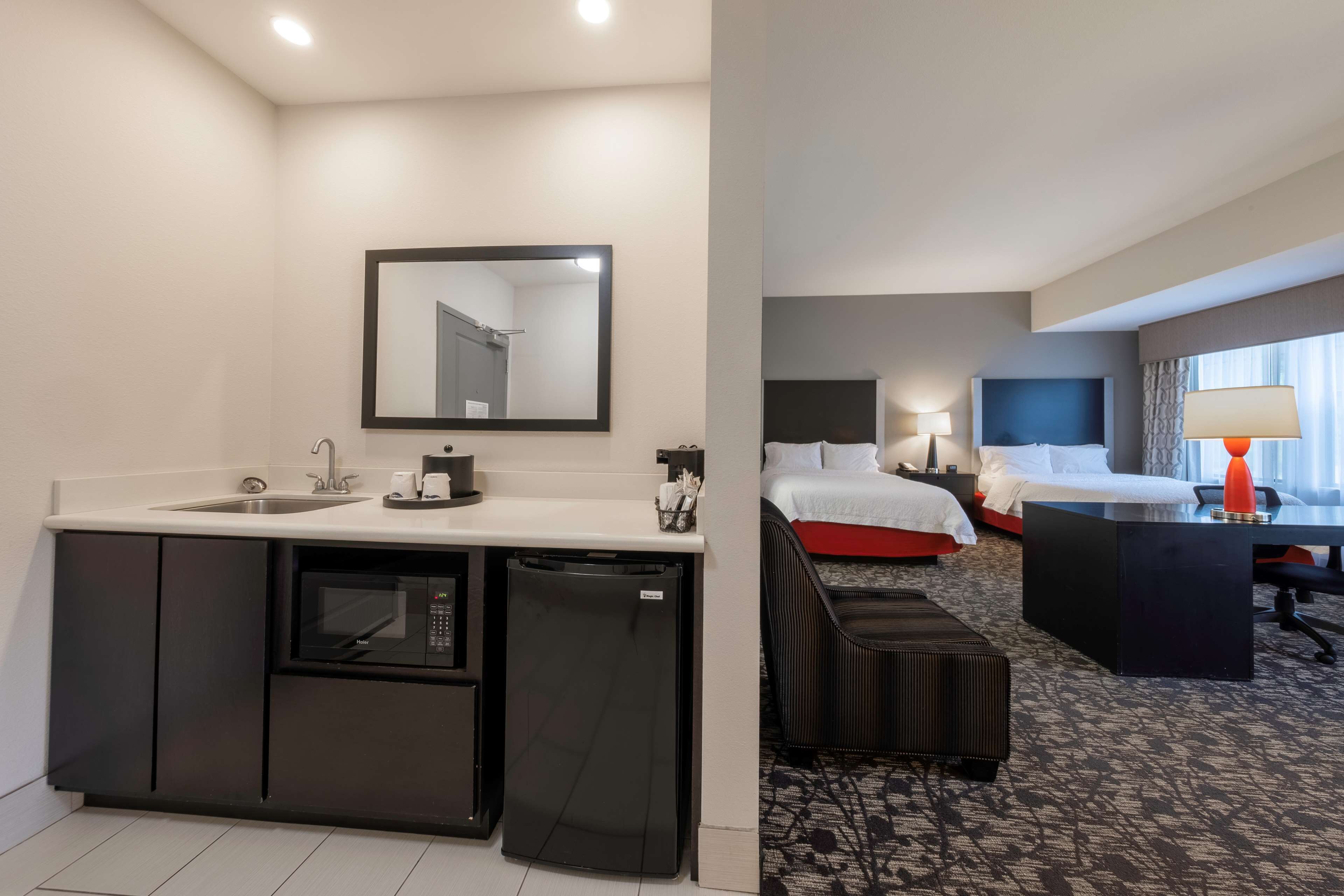 Quarto de hotel com cozinha compacta, cama com roupa de cama vermelha, escrivaninha iluminada por um abajur e um espelho grande.