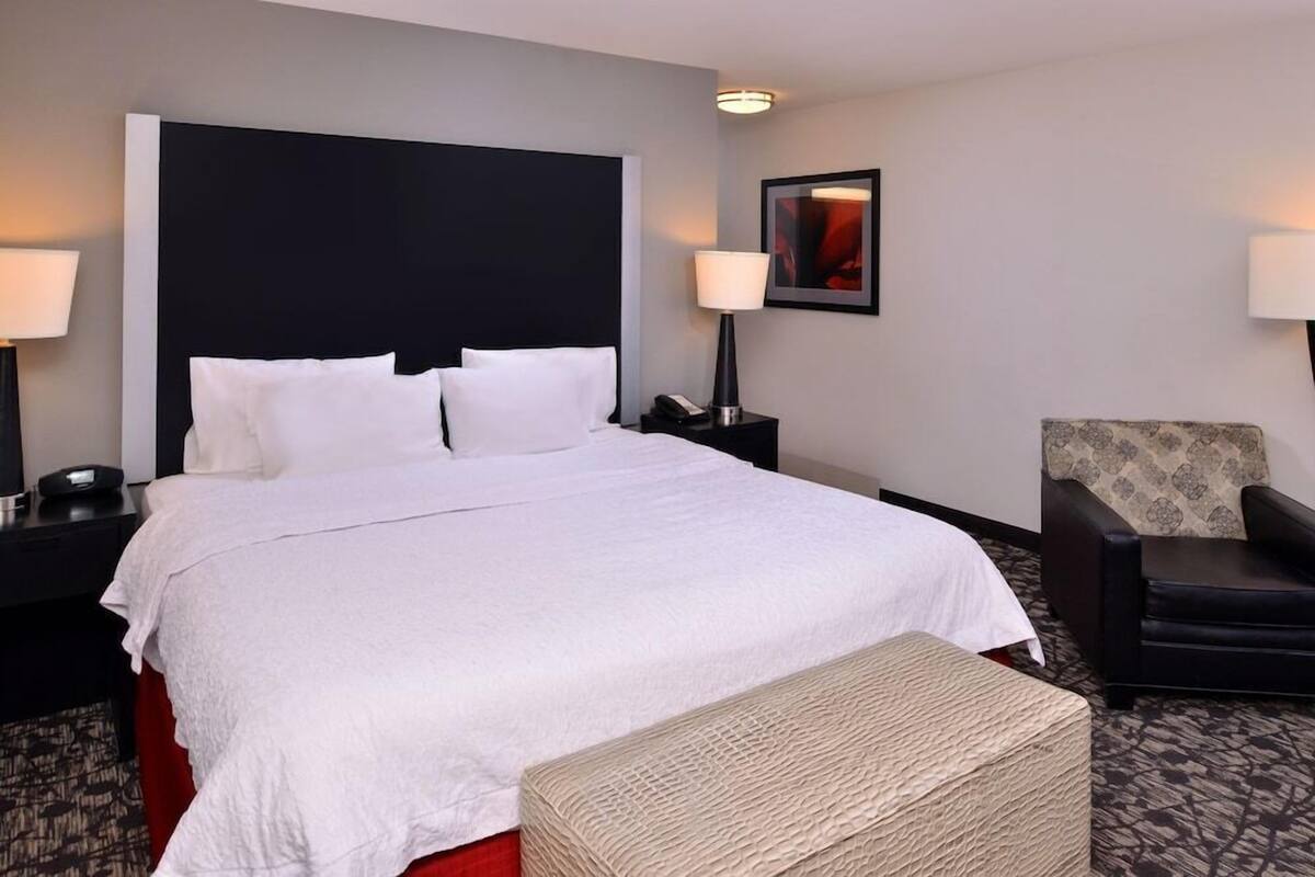 A hotel room with a large bed, a nightstand with a lamp, a chair, and a framed picture on the wall.