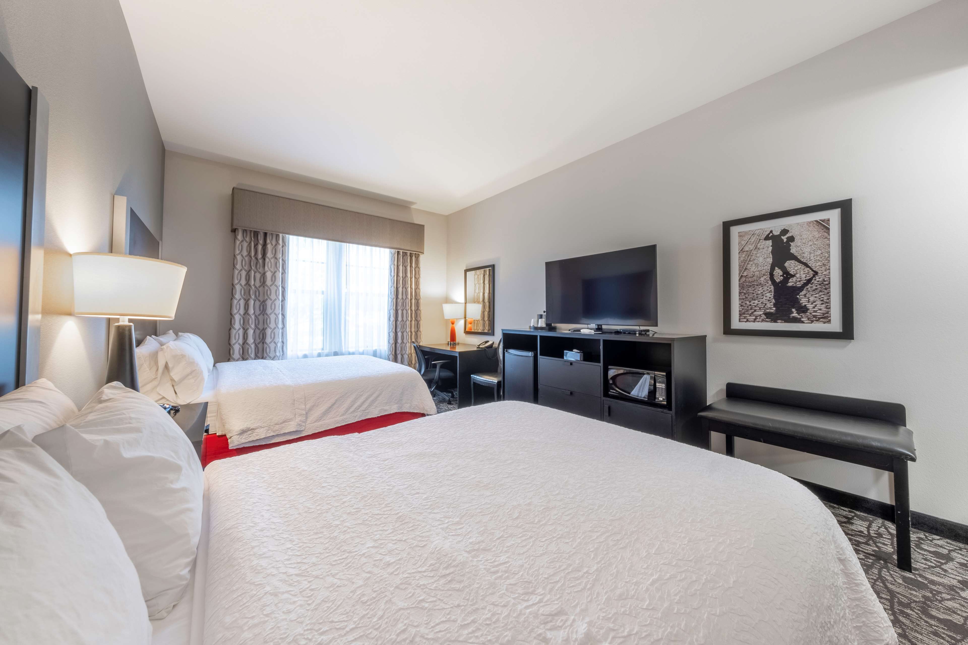 A hotel room with two beds, a desk, a chair, a TV, and a bench.