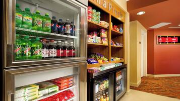 A hotel lobby with a refrigerator stocked with beverages and snacks, a sign reading "In a Pinch," and a wall-mounted sign indicating amenities like "Stay," "Work," and "Play."