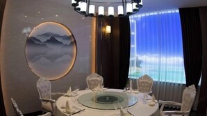 Una mesa redonda de comedor con cubierta de vidrio, rodeada de sillas ornamentales, en una habitación con un espejo circular grande y vistas al mar a través de una ventana con cortinas translúcidas.