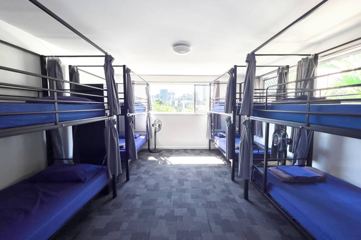 A room with bunk beds, a fan, and a view of buildings through a window.