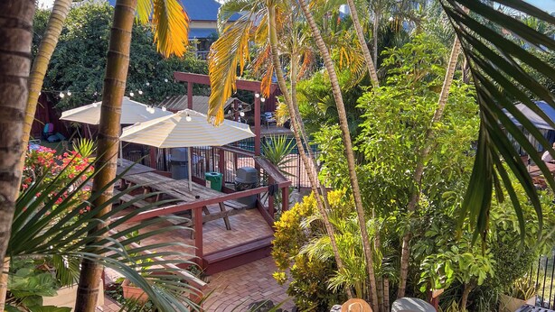 A patio area with a wooden deck, palm trees, and a covered seating area with umbrellas. - Somewhere To Stay Backpackers (Highgate Hill)