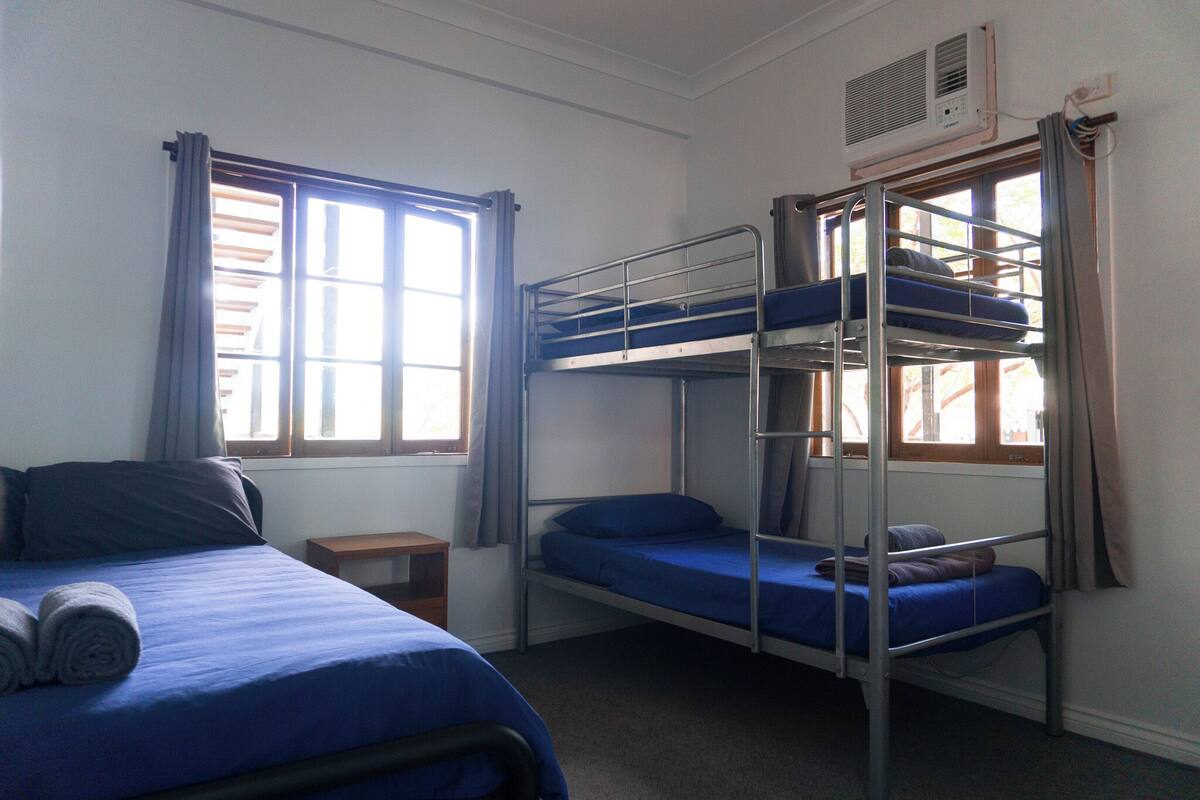 A dormitory room with bunk beds, a small bedside table, and a window with curtains.