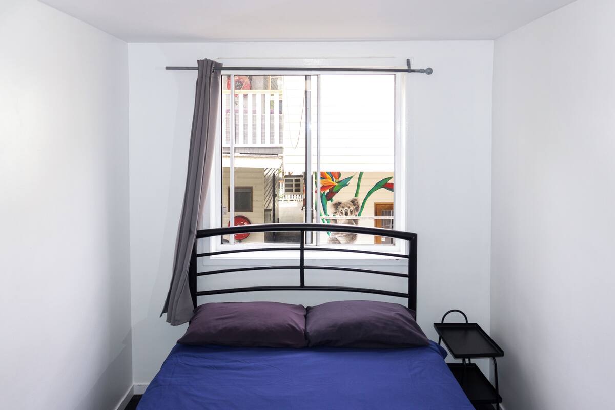 A bedroom with a bed, a window with curtains, a nightstand, and a view of a balcony with a plant.