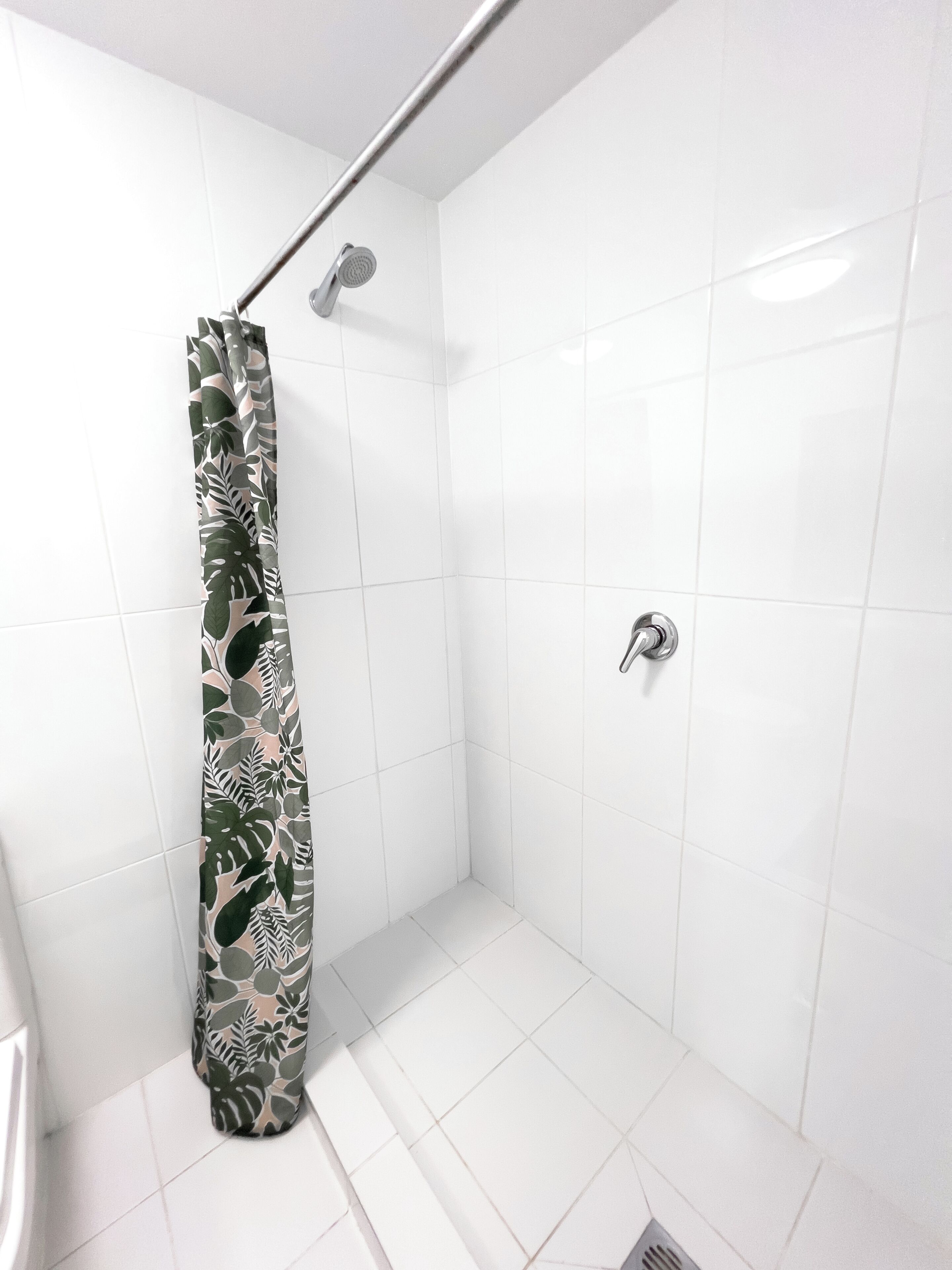 Une petite salle de douche aux murs recouverts de carreaux blancs, avec un rideau de douche orné d’un motif de feuilles.