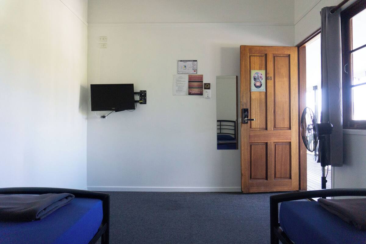 A room with two single beds, a wooden door, a wall-mounted TV, and a fan.