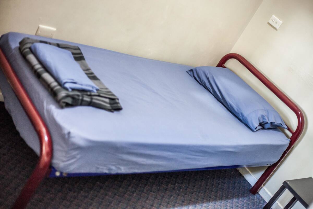 A single bed with a blue mattress and a striped blanket, a red metal frame, and a small table beside it.