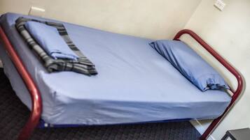 Ein Einzelbett mit blauer Matratze und einer gestreiften Decke, einem roten Metallgestell und einem kleinen Tisch daneben.