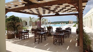 Un patio al aire libre con mesas y sillas de madera, una piscina al fondo y vista al océano.