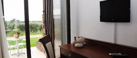 A room with a wooden desk, a chair, a television mounted on the wall, and a sliding glass door leading to an outdoor area with a table and chairs.