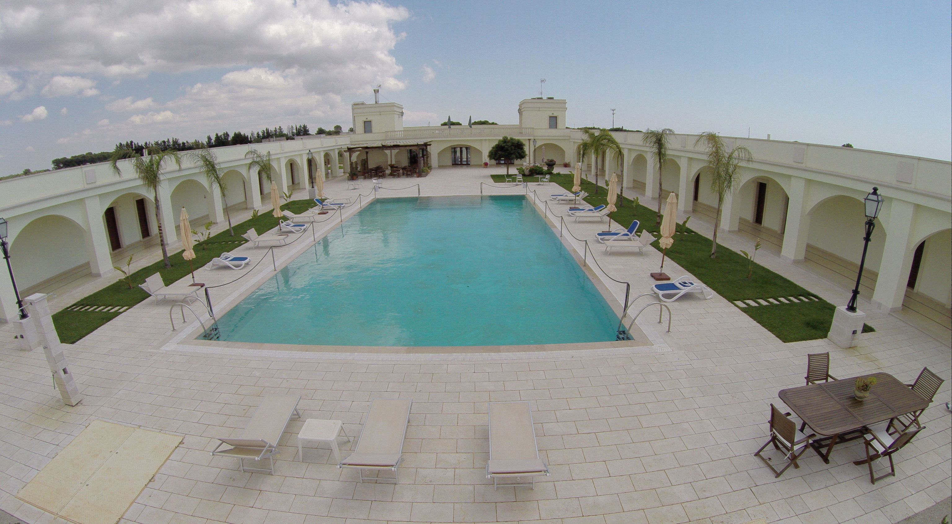 A rectangular pool with a clear blue water surface, surrounded by white tiled flooring and a series of white lounge chairs and umbrellas.