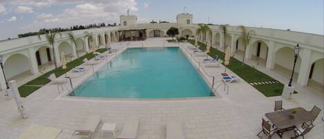 A rectangular pool with a clear blue water surface, surrounded by white tiled flooring and a series of white lounge chairs and umbrellas.