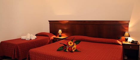 Quarto de hotel com duas camas de solteiro e uma cama maior, cada uma com roupa de cama vermelha e cabeceira de madeira. Há luminárias de cabeceira e uma pequena mesa com um abajur.