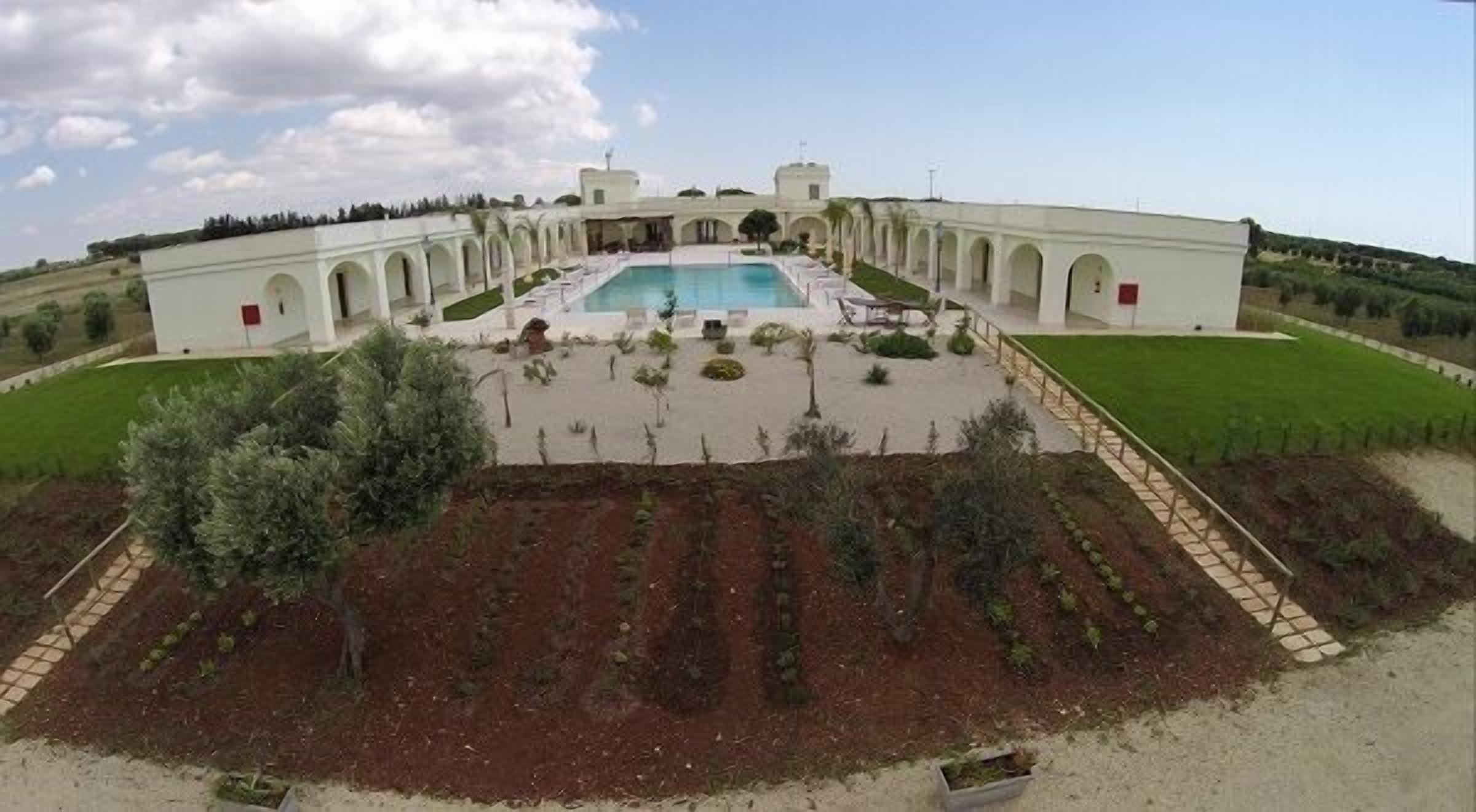 A large building with a central pool, surrounded by a garden with rows of plants and a few trees.
