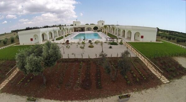 A large building with a central pool, surrounded by a garden with rows of plants and a few trees.