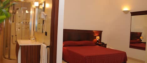 A hotel room with a shower, a sink, a bed, and a nightstand with a lamp.