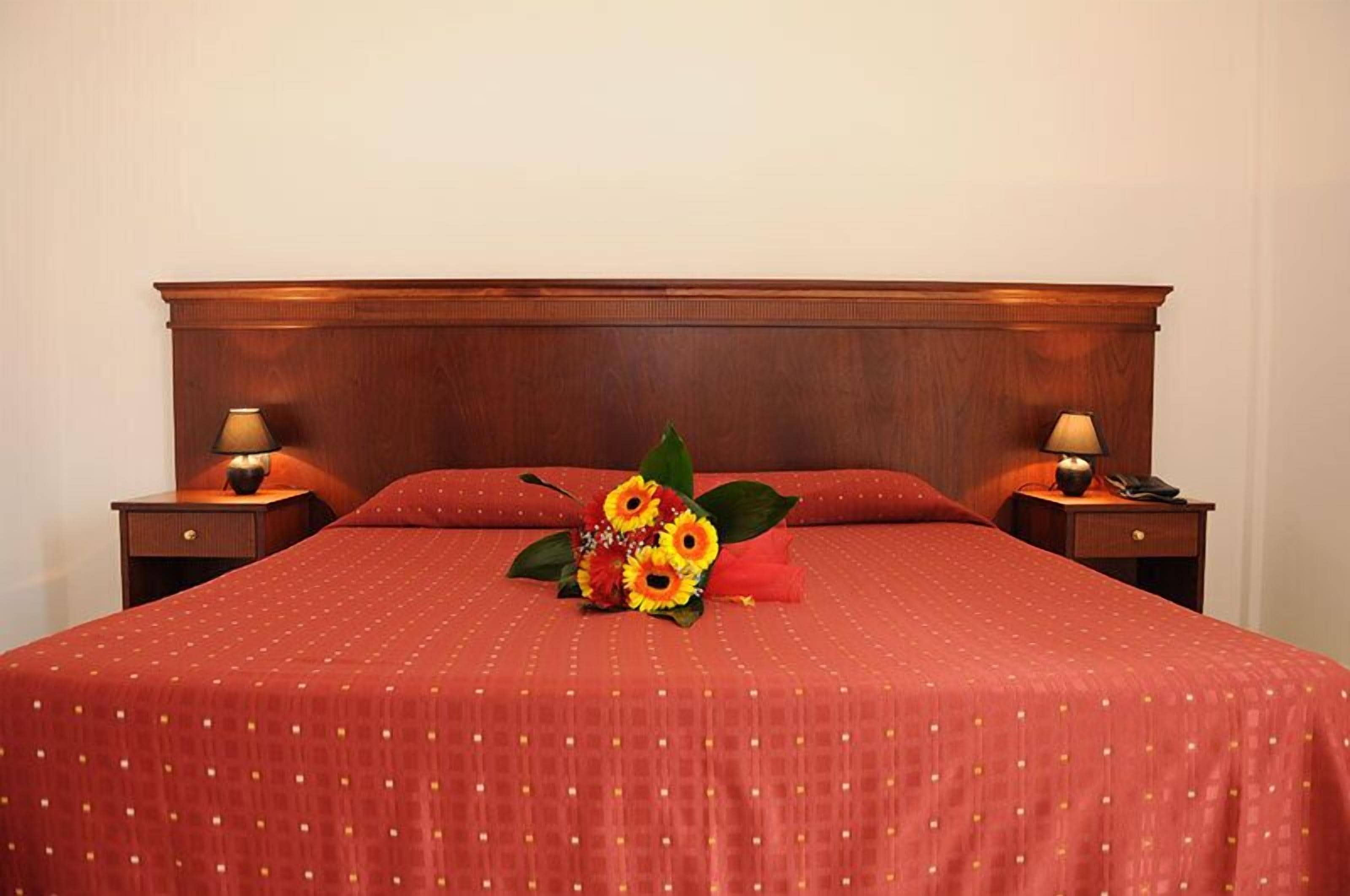 A neatly made bed with a red bedspread and a bouquet of sunflowers. There are two bedside tables with lamps on either side of the bed.
