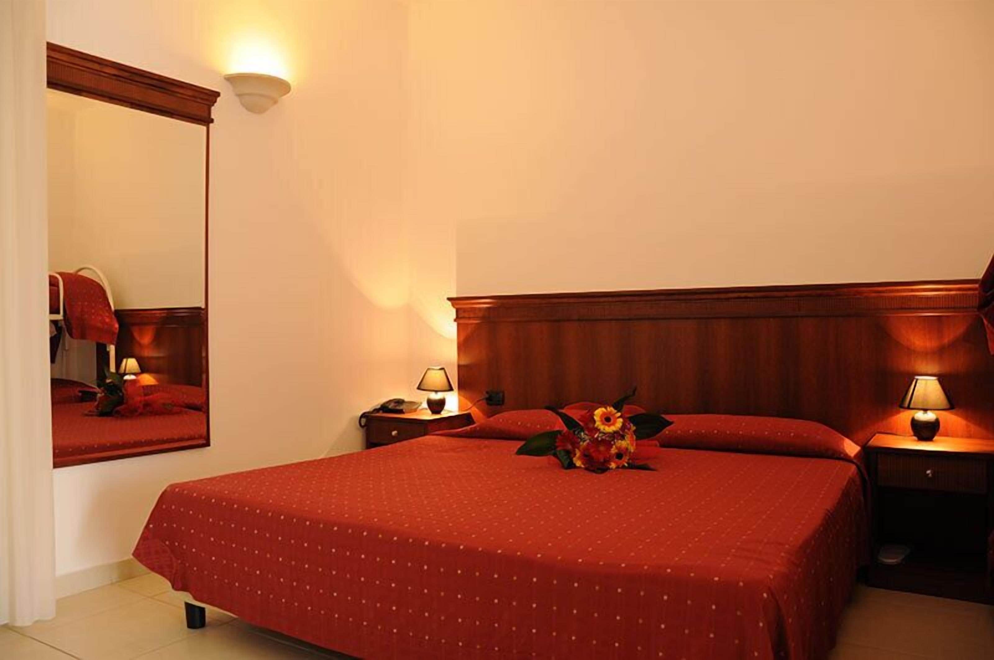 A hotel room with a large bed, two bedside tables with lamps, a mirror, and a red floral arrangement on the bed.