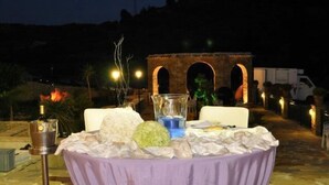 An outdoor dining table set up with a purple tablecloth, surrounded by chairs, with a stone floor and a building in the background. - Casale del Golfo (Alcamo)