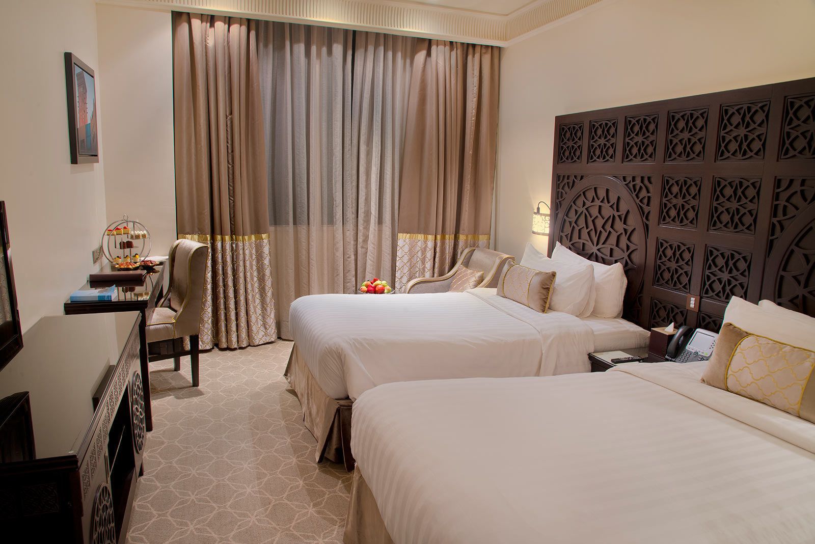 A hotel room with two beds, a desk with a chair, a decorative headboard, and a large window with curtains.