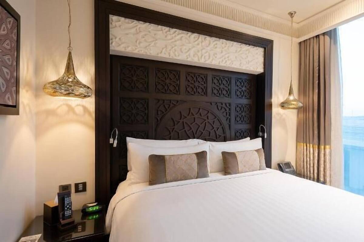 A hotel room with a large bed, a decorative headboard, a bedside table with a lamp, and a view of the city through the window.