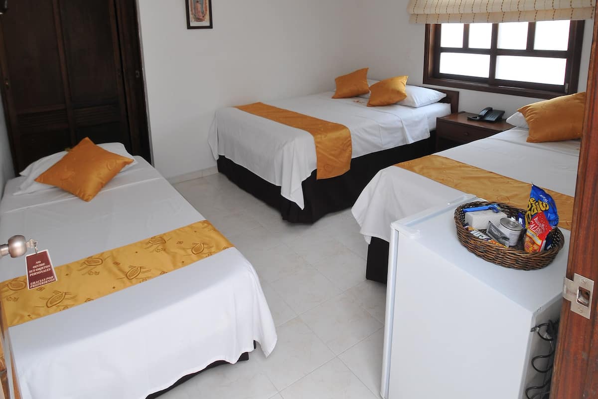 Habitación de hotel con tres camas, una mesita pequeña, una ventana con persianas, una lámpara en el techo y un armario de madera.