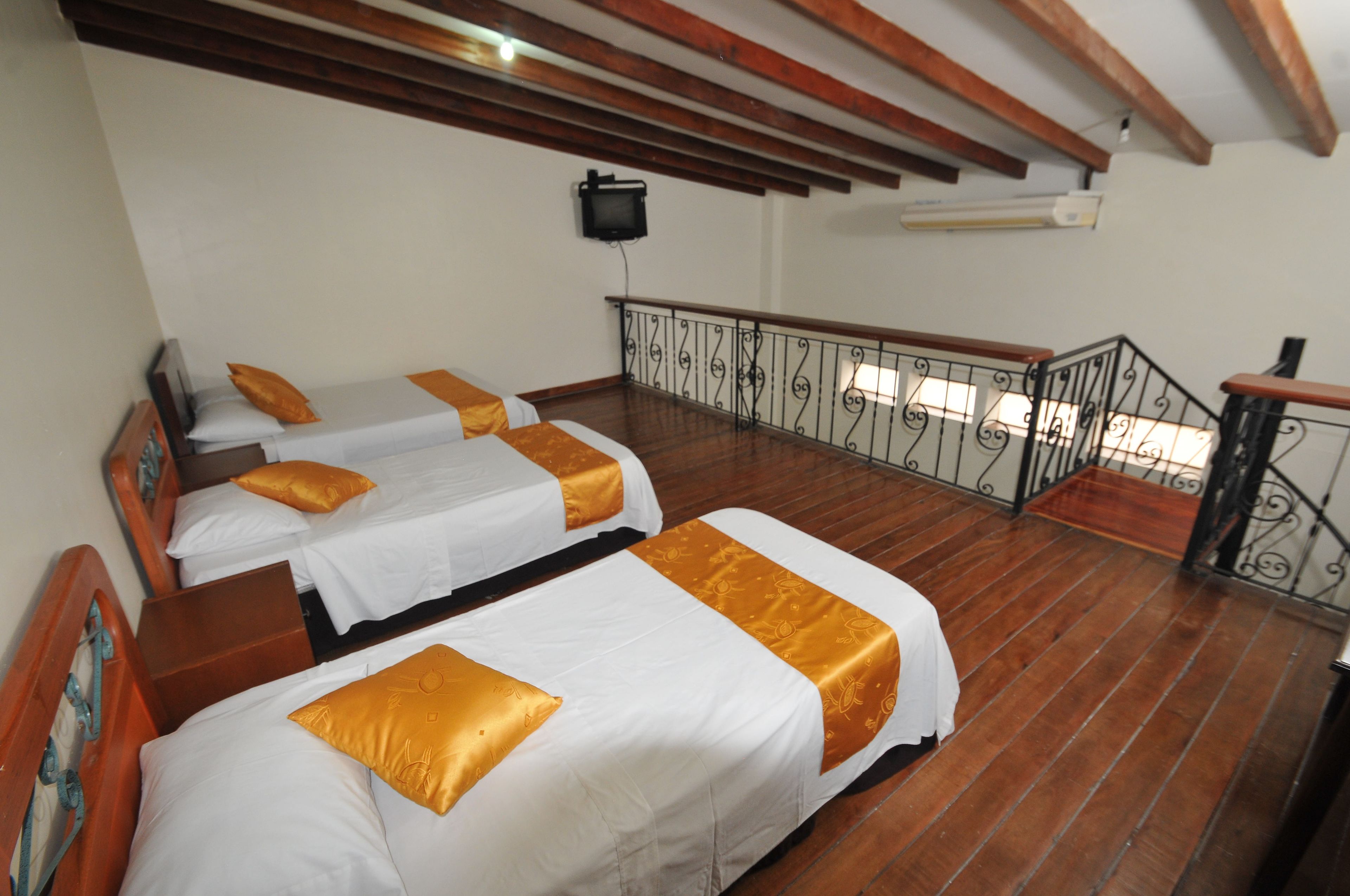 A hotel room with three beds, wooden flooring, a balcony with a metal railing, and a television mounted on the wall.