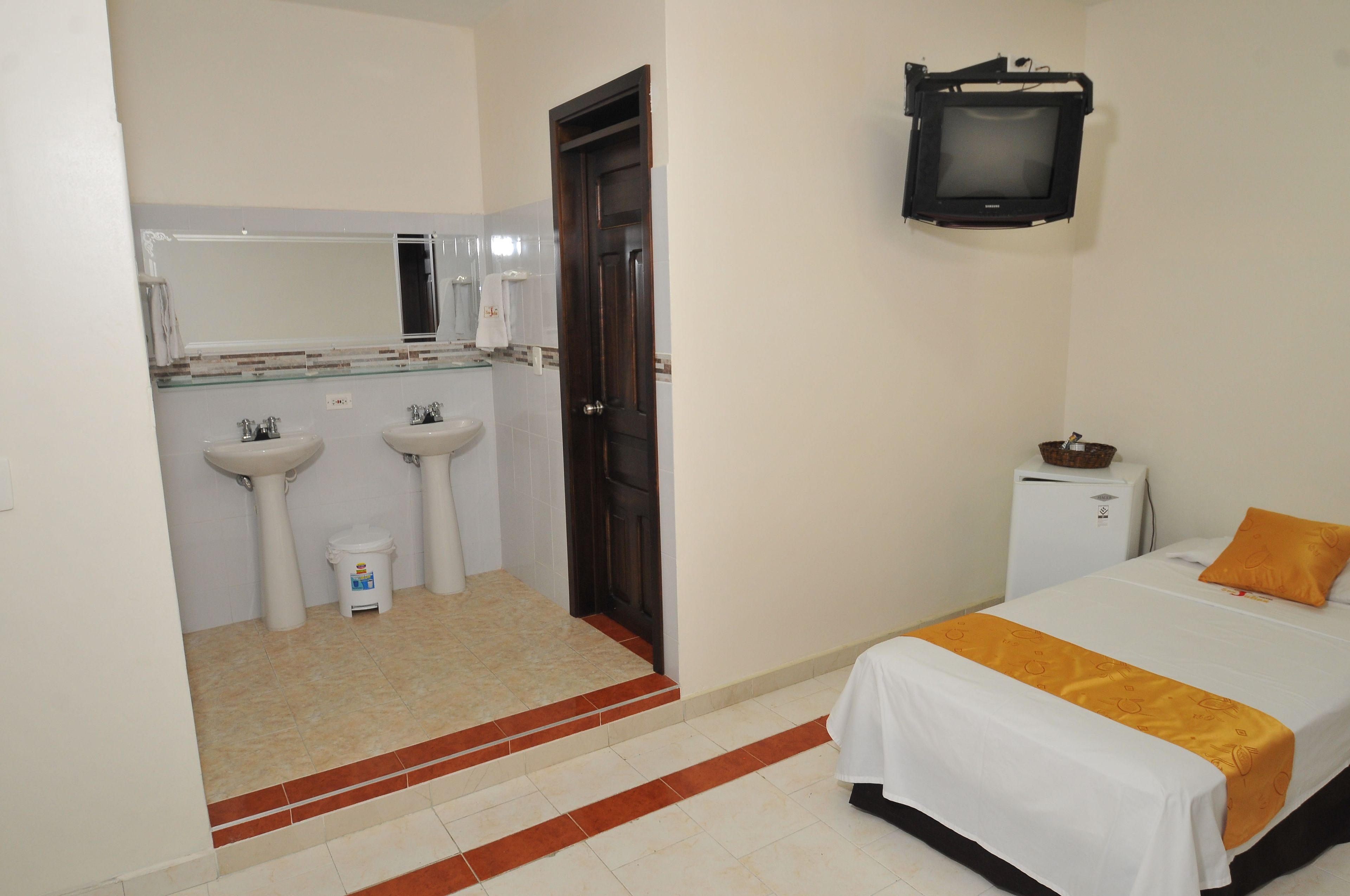Una habitación de hotel con una cama, un televisor montado en la pared y dos lavamanos en el baño.