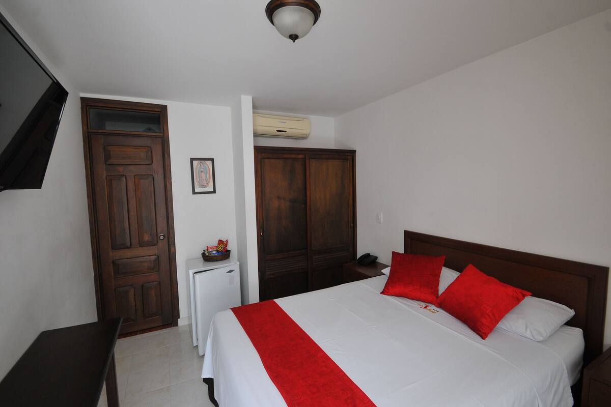 Un dormitorio con cama, un armario de madera, una mesita con una cesta y un televisor montado en la pared.