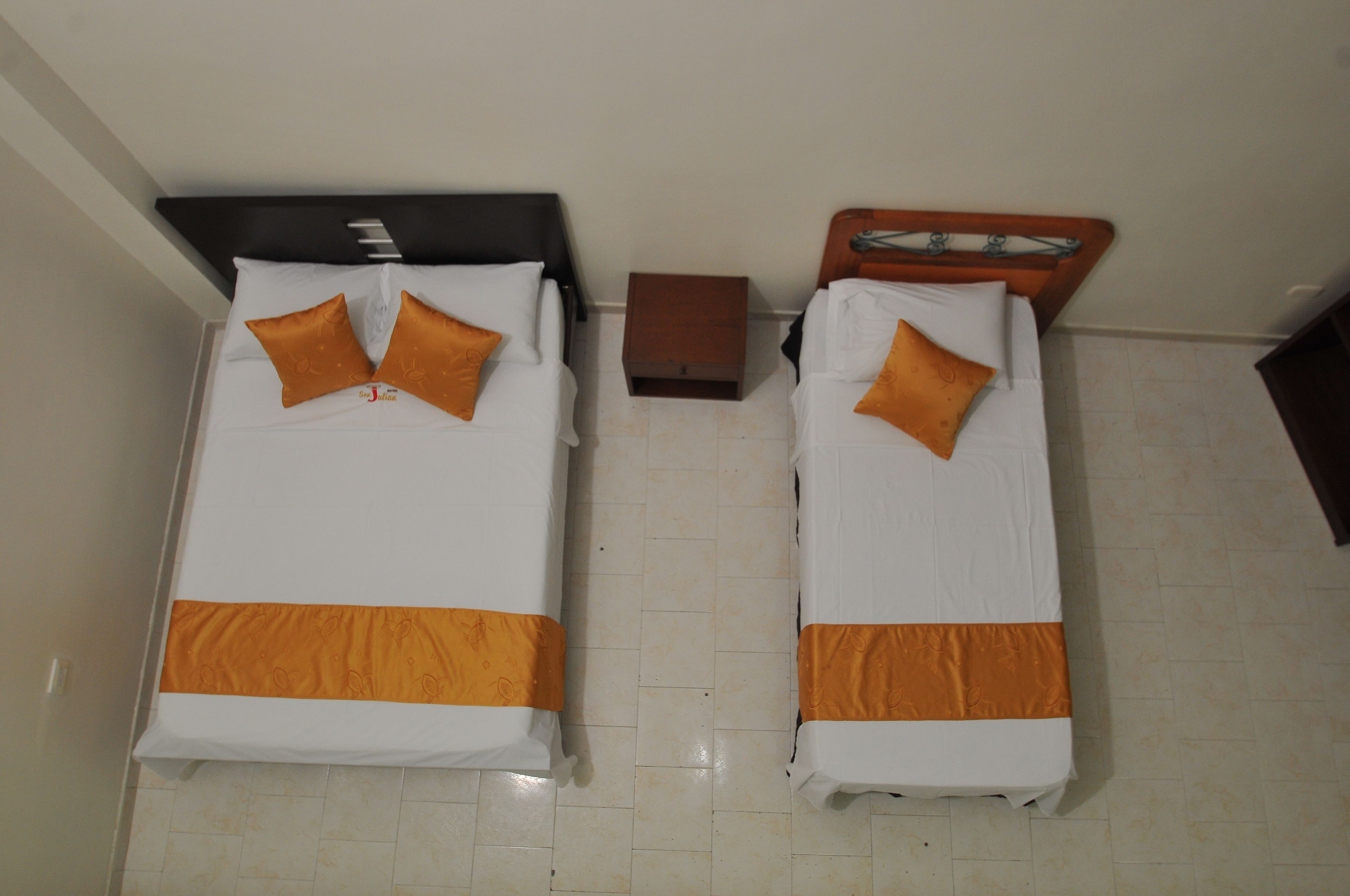 Two single beds with white and orange bedding, a small wooden nightstand, and a tiled floor.