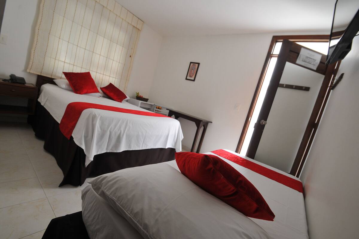 Habitación de hotel con dos camas, cada una con ropa de cama blanca y detalles rojos. Hay un escritorio con una silla, una mesita con una lámpara y una ventana con persianas.