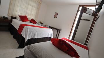Habitación de hotel con dos camas, cada una con ropa de cama blanca y detalles rojos. Hay un escritorio con una silla, una mesita con una lámpara y una ventana con persianas.