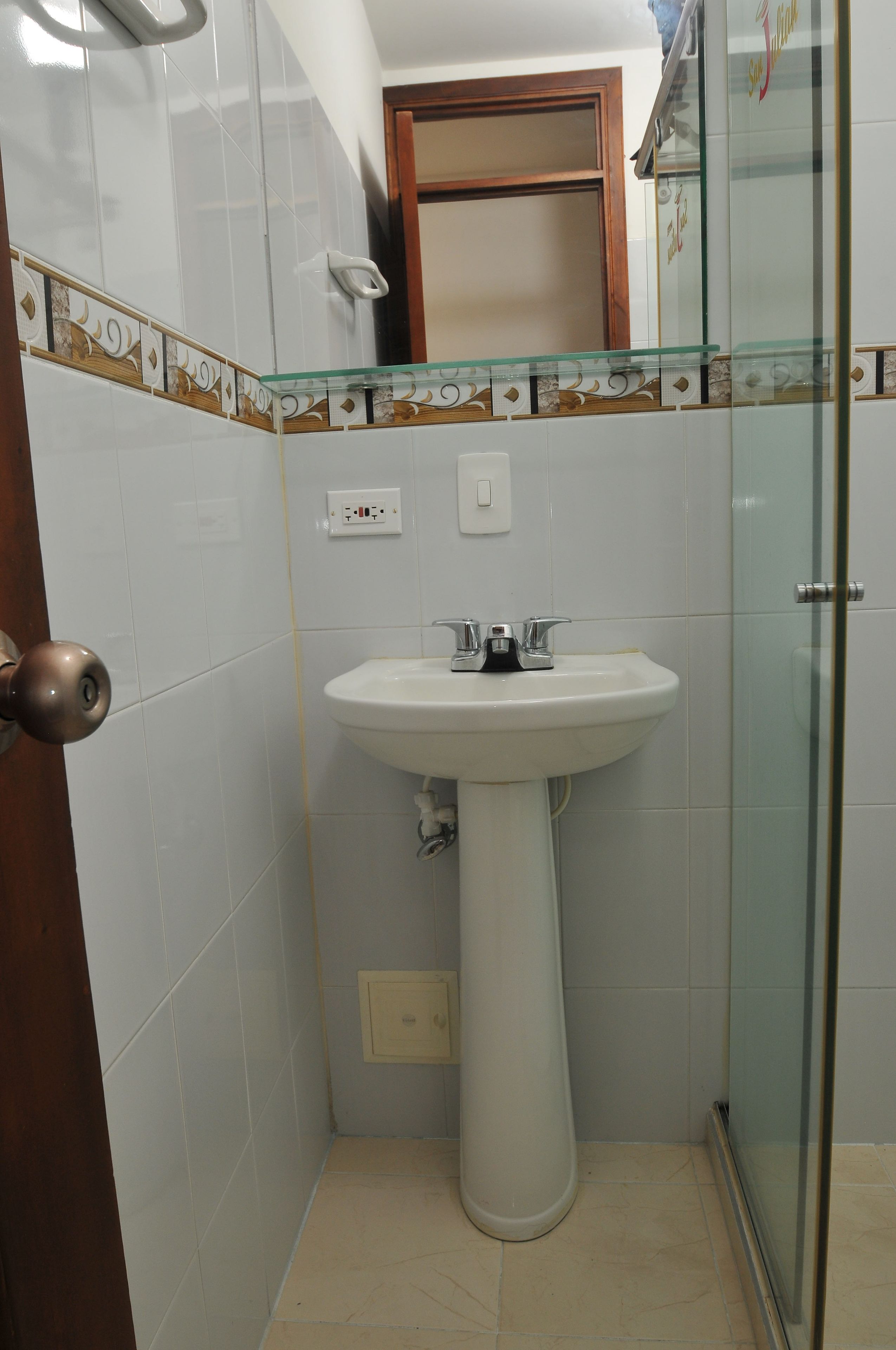 Un baño con lavamanos pedestal, cabina de ducha con mampara de vidrio y puerta de madera.