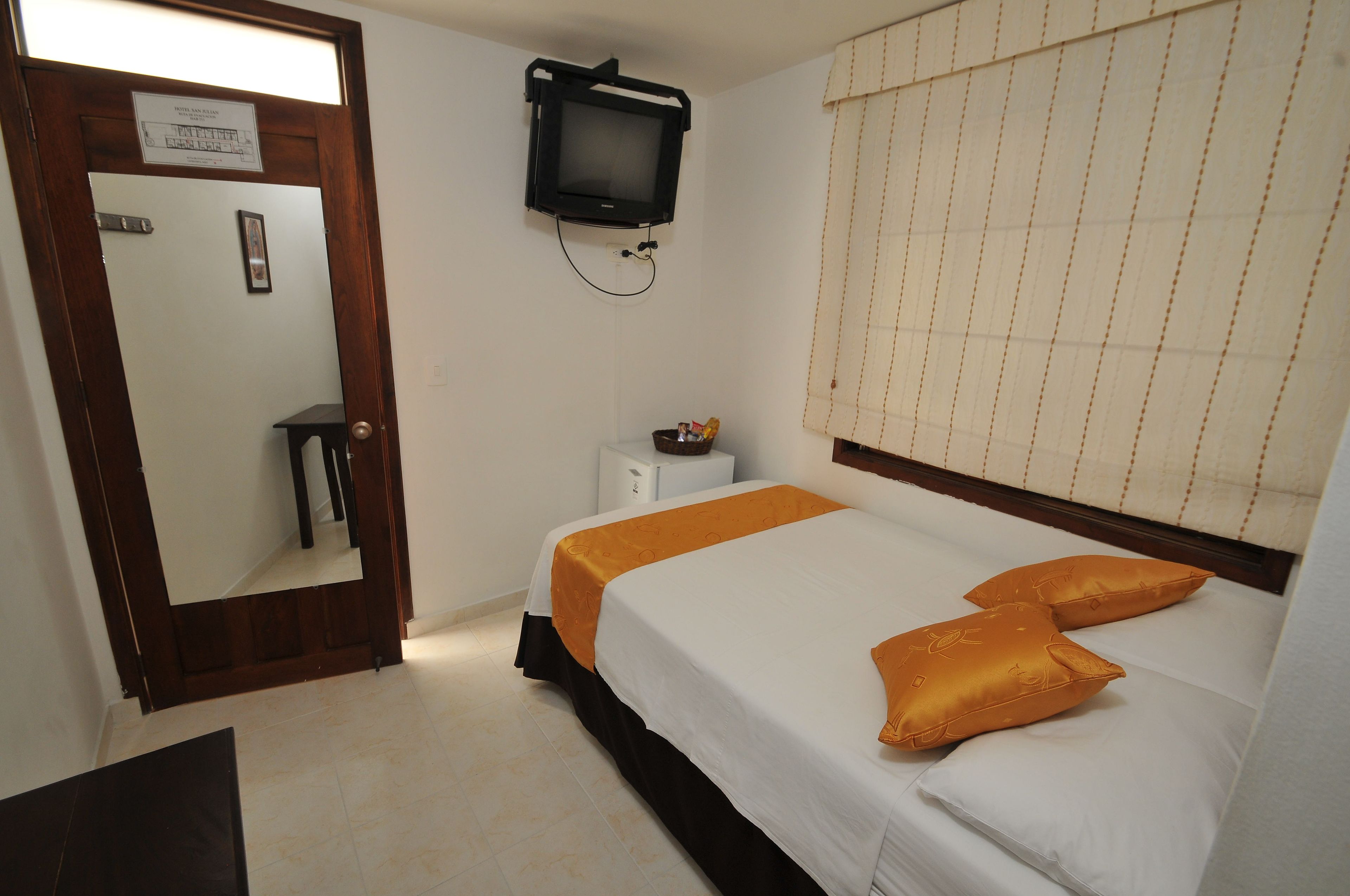 A hotel room with a bed, a television mounted on the wall, a small table, and a window with blinds.