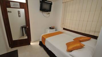 A hotel room with a bed, a television mounted on the wall, a small table, and a window with blinds.