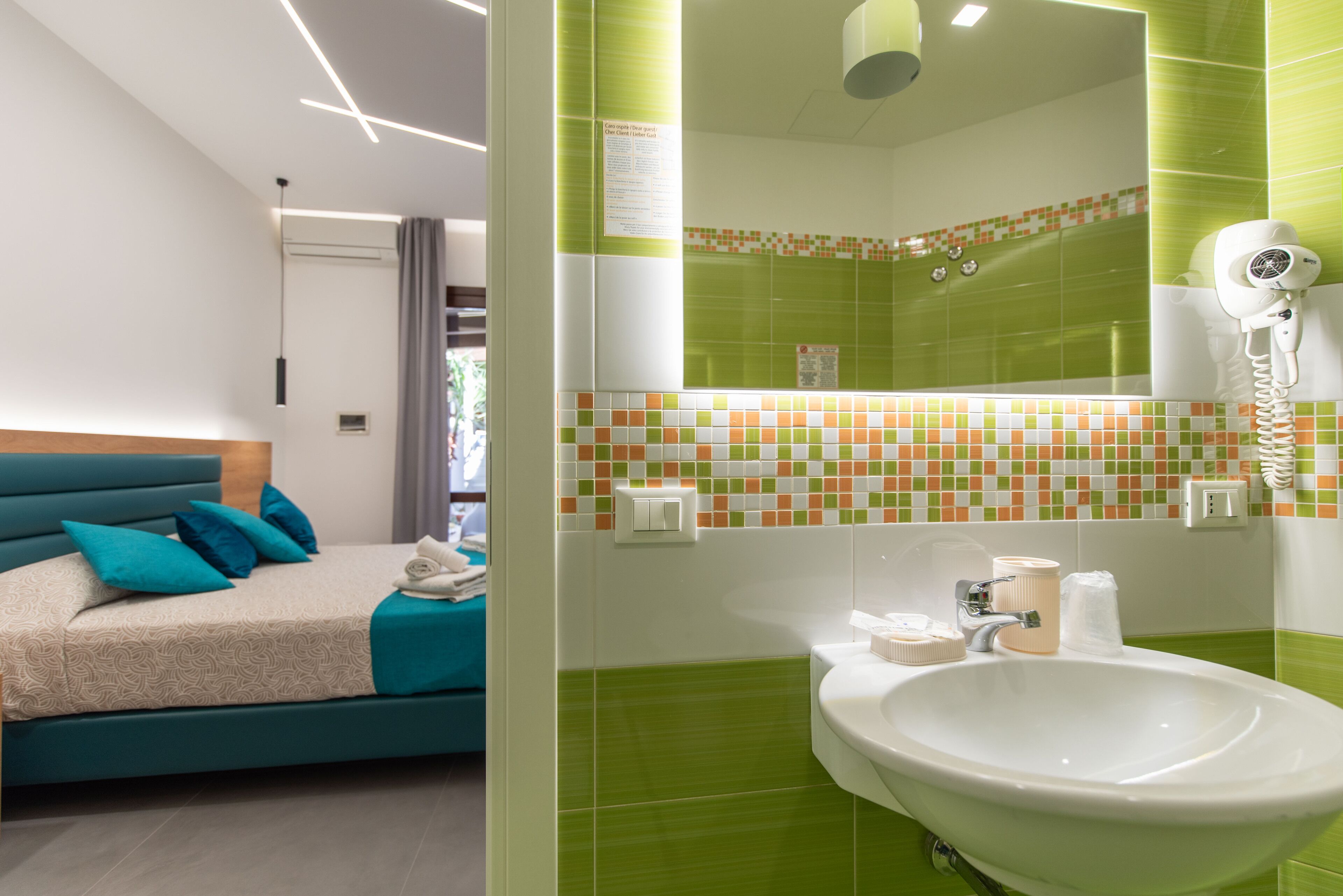 Una habitación de hotel moderna con una cama color turquesa, un lavamanos blanco grande y una zona de ducha con azulejos verdes.
